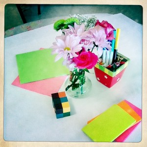 Flowers in a vase surrounded by pens and brightly colored pieces of paper and notes
